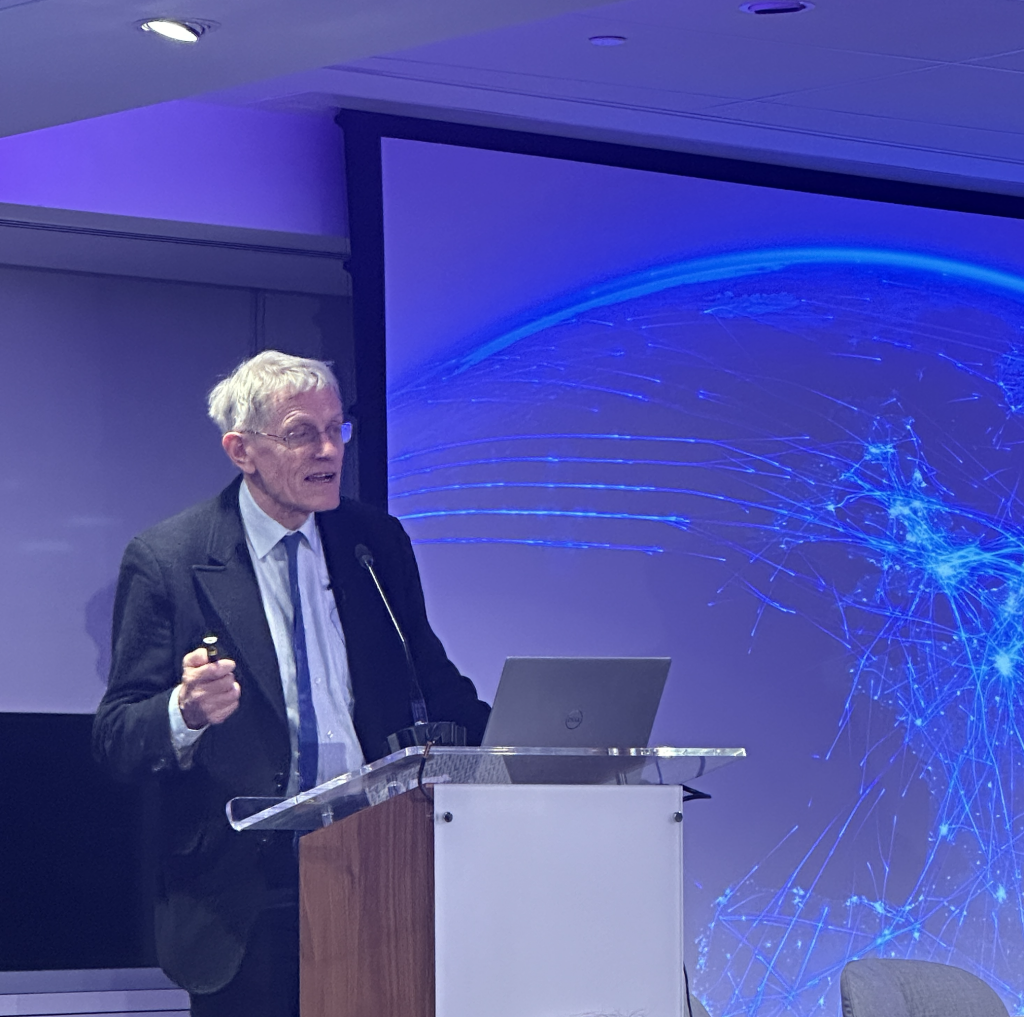 Simon Calder, travel journalist, speaking at a podium against a blue screen showing aeroplane flight paths across the globe, at the Association of Serviced Apartment Providers Annual Conference, 2024